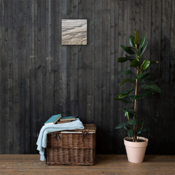 Una cesta de mimbre con libros y una manta doblada reposa contra una pared de madera oscura, mientras que el "Lienzo surcos arena mojada" añade encanto en la parte superior. A la derecha, una maceta alta completa la escena con un toque elegante.