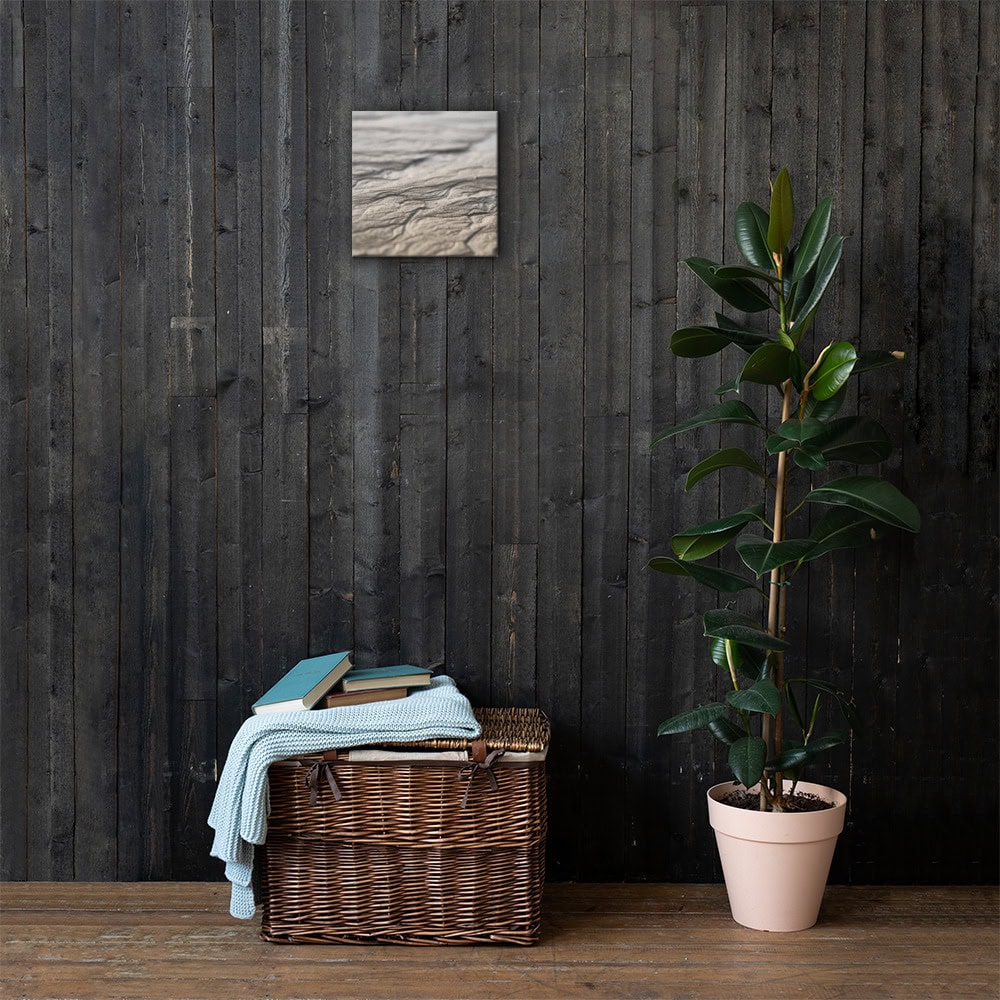 Una cesta de mimbre con libros y una manta doblada reposa contra una pared de madera oscura, mientras que el "Lienzo surcos arena mojada" añade encanto en la parte superior. A la derecha, una maceta alta completa la escena con un toque elegante.