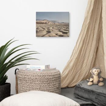 Un acogedor rincón de la habitación cuenta con una cortina beige, una pila de cojines y un peluche. Un puf redondo con textura sostiene varios libros de Tienda Online. Cerca hay una planta en maceta. Adornando la pared blanca está "Lienzo Sotavento", una cautivadora fotografía de un paisaje desértico de Volcanica Productions.