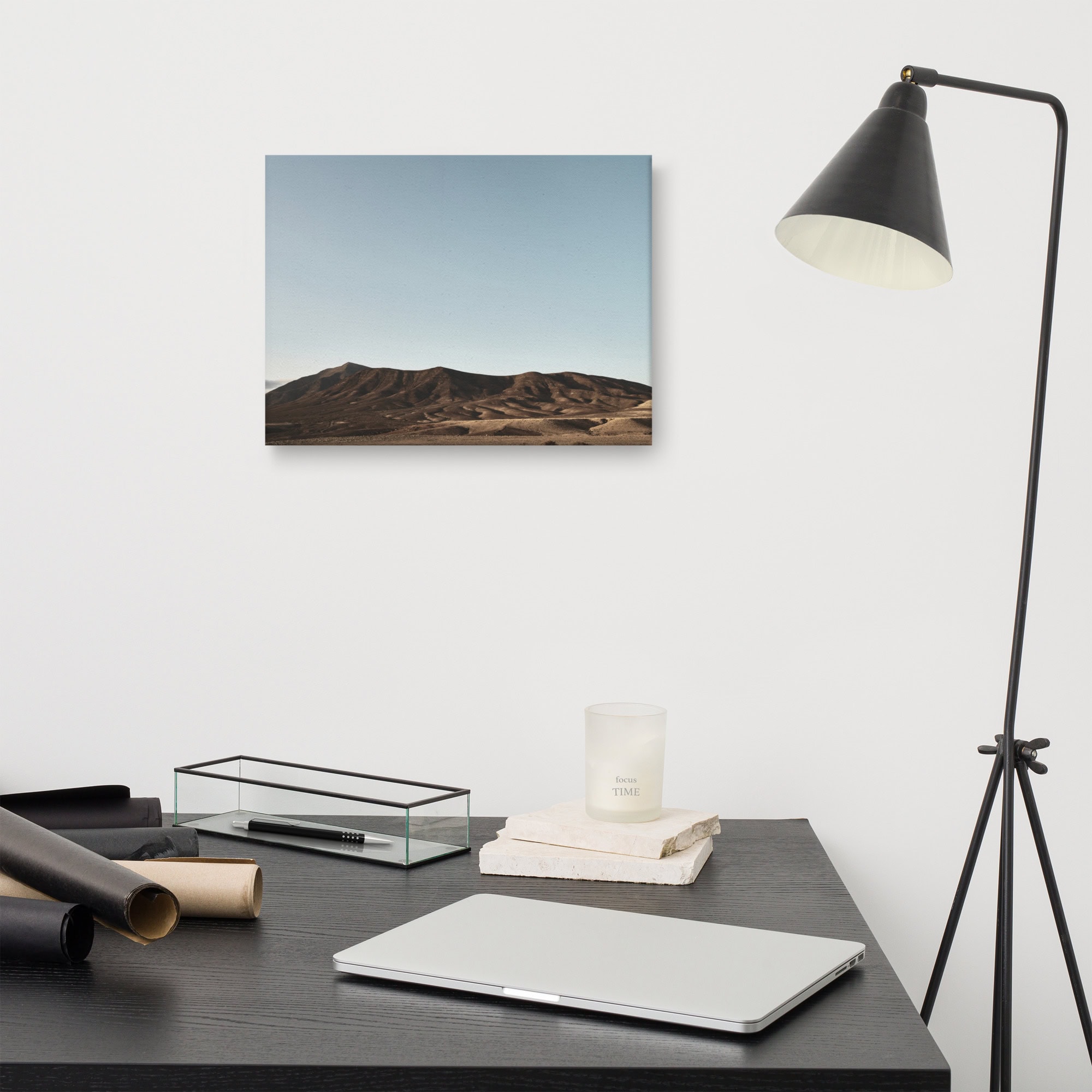 Un espacio de trabajo minimalista con un escritorio negro con una computadora portátil, elementos decorativos y una lámpara de pie negra se complementa con el encanto vibrante de la fotografía del paisaje enmarcado "Lienzo Los Ajaches" colgada en la pared blanca, todo bajo un cielo despejado.