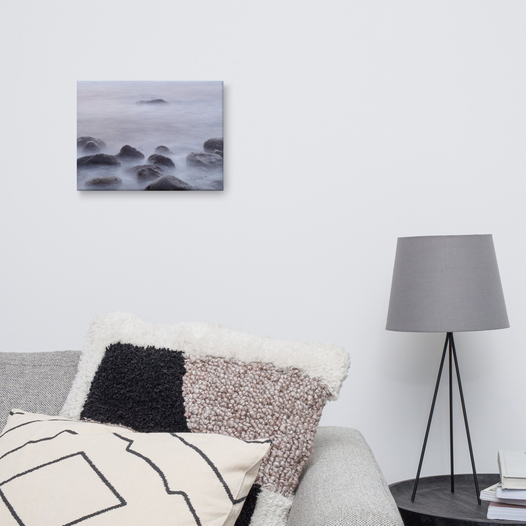 Una sala de estar minimalista muestra un sofá gris adornado con almohadas texturizadas y una moderna lámpara de trípode negra. Colgado en la pared blanca de arriba se encuentra el "Lienzo de Rocas de la playa de El Lloret", una pintura de paisaje marino que captura la belleza agreste de La Palma. Junto al sofá, una mesa auxiliar negra contiene libros, lo que realza la atmósfera serena.