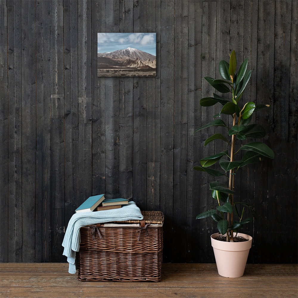 Una cesta de mimbre con libros y una manta azul claro reposa sobre una pared de madera oscura, acompañada de una planta verde en maceta a la derecha. Arriba, la foto del paisaje del Lienzo Teide de Arte Decorativo añade elegancia a la escena.