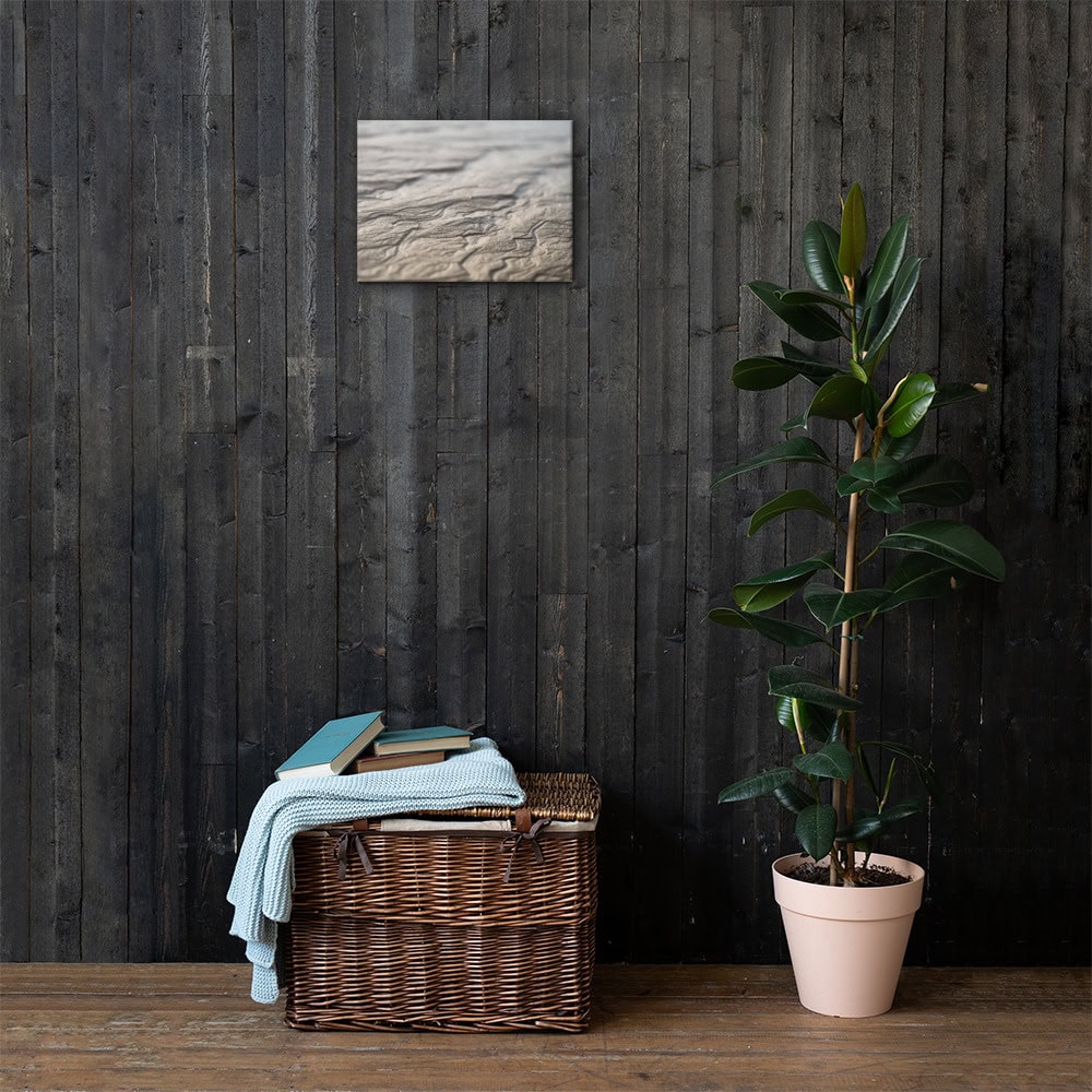 Una cesta de mimbre llena de libros y una manta azul se encuentra junto a una maceta sobre un suelo de madera, capturando la esencia tranquila de los paisajes de La Palma. Cerca de allí, "Lienzo surcos arena mojada" decora la pared de madera oscura, ofreciendo un elemento artístico que refleja el encanto de un descubrimiento artesanal de Tienda Online.