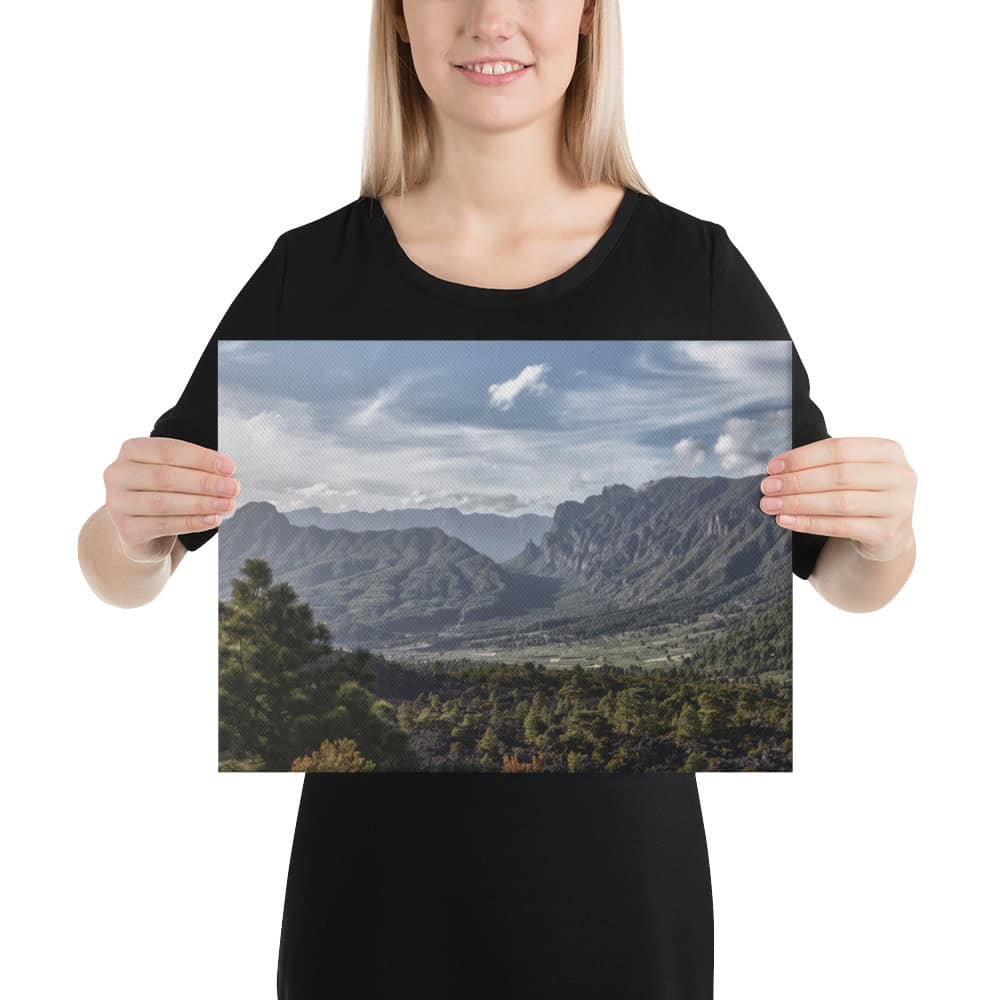 Una persona con una camisa negra sostiene el Lienzo Caldera de Taburiente de Volcanica Productions, un impresionante paisaje que representa montañas, un valle y un cielo nublado. Están colocados sobre un sencillo fondo blanco, fusionando el arte con la elegancia de la naturaleza.