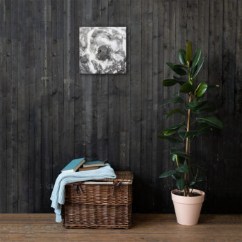 Una cesta de mimbre llena de libros y una manta azul doblada se apoya contra una pared revestida de madera oscura, capturando a la perfección la esencia de Arte Decorativo. Sobre ella se encuentra "Lienzo roca y sal", una pintura abstracta en escala de grises. A la derecha, una planta en maceta con hojas de color verde oscuro de Paisajes Canarios contribuye a crear una atmósfera serena y artística a la vez.