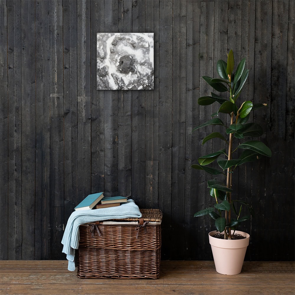 Una cesta de mimbre llena de libros y una manta azul doblada se apoya contra una pared revestida de madera oscura, capturando a la perfección la esencia de Arte Decorativo. Sobre ella se encuentra "Lienzo roca y sal", una pintura abstracta en escala de grises. A la derecha, una planta en maceta con hojas de color verde oscuro de Paisajes Canarios contribuye a crear una atmósfera serena y artística a la vez.