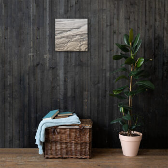 Una habitación rústica con una pared de madera oscura muestra el pequeño cuadro titulado "Lienzo surcos arena mojada". A la derecha hay una planta frondosa en una maceta, mientras que a la izquierda, una cesta de mimbre contiene libros y una manta azul doblada, posiblemente adquirida en una tienda en línea inspirada en el encanto de La Palma.