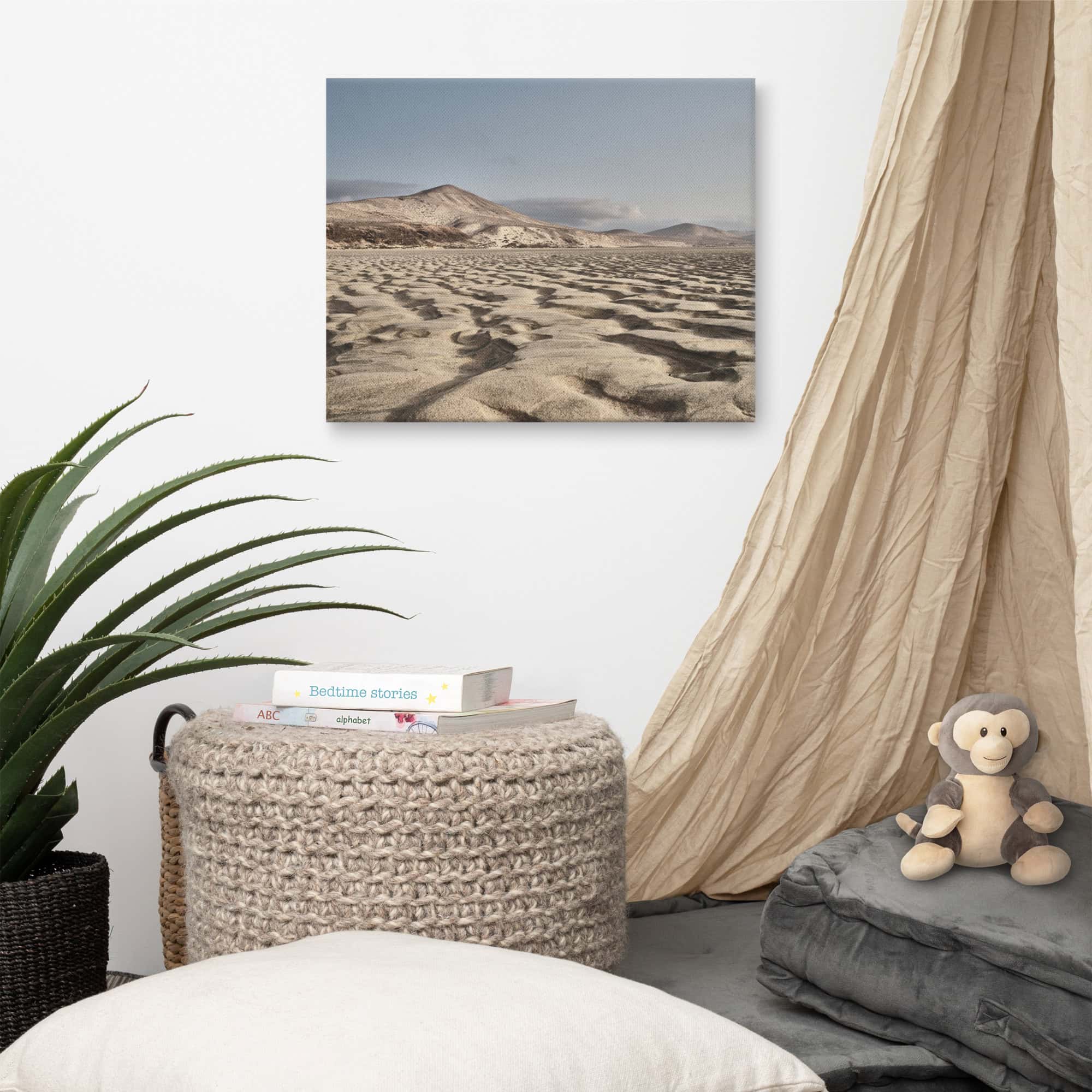 Un rincón acogedor cuenta con una otomana de nudos, una pila de libros y un peluche que reposa sobre cojines grises. Una cortina beige cuelga del lado derecho. Arriba, la pared blanca está adornada con el cuadro de paisaje "Lienzo Sotavento", que representa un terreno rocoso y montañas que recuerdan a Paisajes Canarios.