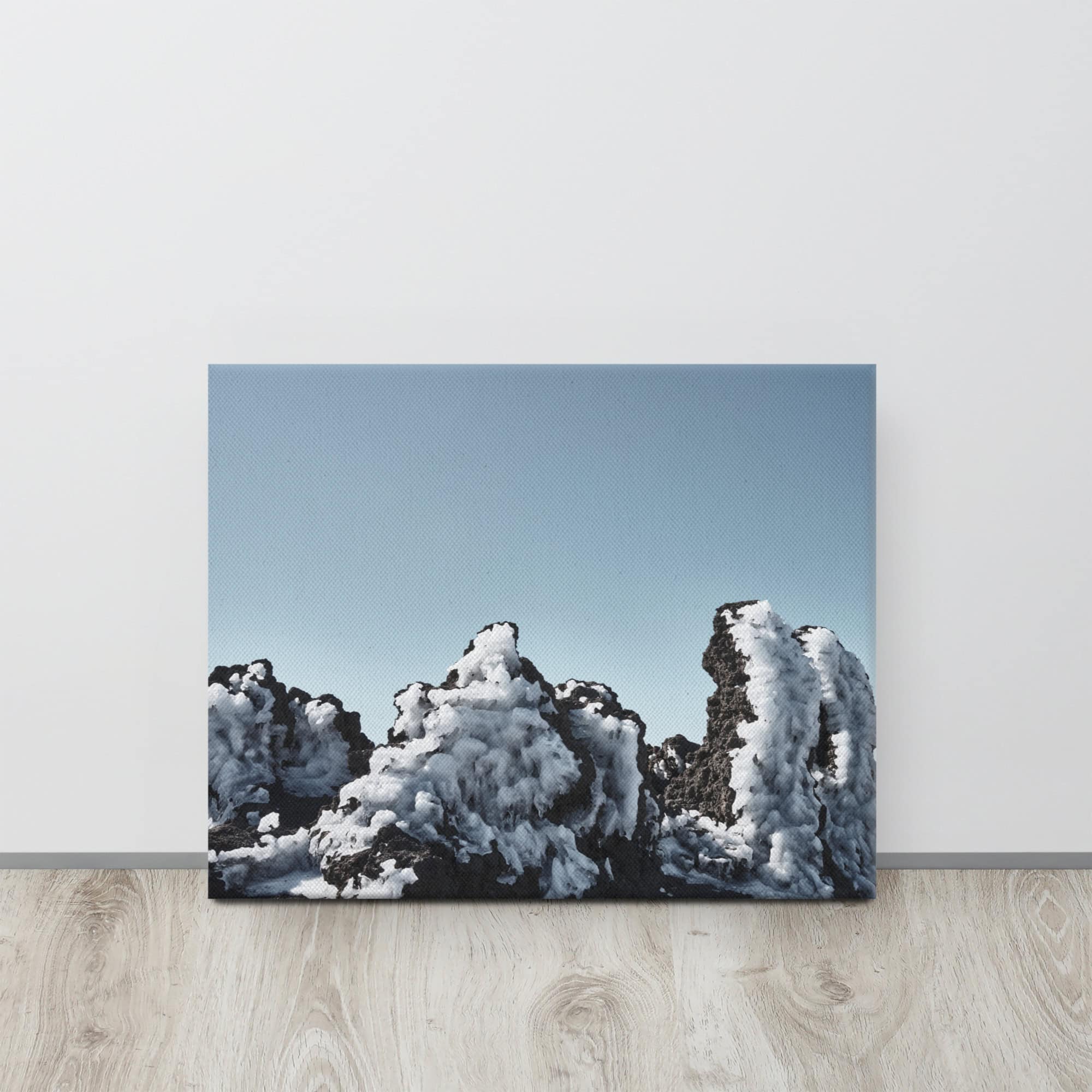 Un lienzo titulado "Lienzo Roque helado", apoyado sobre una pared blanca, muestra un paisaje de formaciones rocosas negras cubiertas de nieve bajo un cielo azul claro. Esta escena tranquila, en contraste con la calidez del suelo de madera, encapsula la belleza distintiva que se encuentra en Paisajes Canarios, similar a Fotografías impresas de Volcanica Productions.