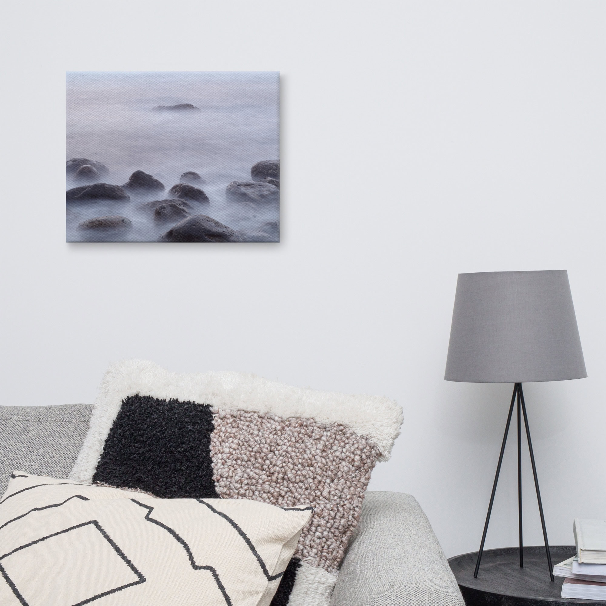 Una tranquila sala de estar con un sofá gris y cojines estampados se encuentra junto a una mesa redonda adornada con libros y una lámpara gris. Colgado en la pared blanca sobre el sofá se encuentra el sereno "Lienzo de Rocas de la playa de El Lloret" de Volcanica Productions, que representa rocas en las brumosas aguas de La Palma.