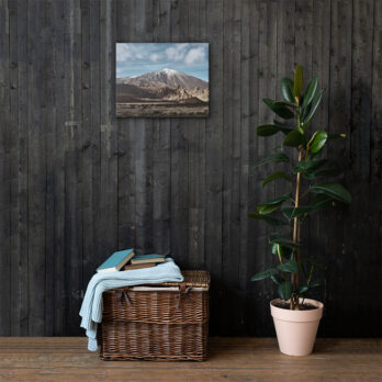 En una habitación rústica, una cesta de mimbre que contiene mantas dobladas y libros se encuentra junto a una planta verde en maceta. Un cuadro de paisaje titulado "Lienzo Teide" adorna la pared de madera oscura, realzando el ambiente acogedor. Esta combinación de Arte Decorativo introduce la tranquila belleza de la naturaleza en el interior.