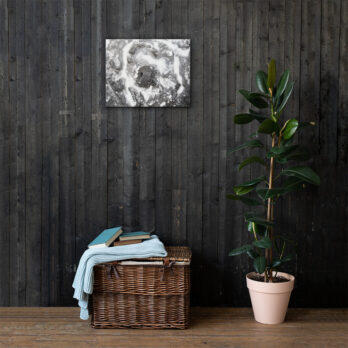 Una cesta de mimbre con libros y una manta doblada reposa sobre un suelo de madera contra una pared revestida de madera oscura, representando el Arte Decorativo. A la derecha se encuentra una planta en maceta con hojas verdes brillantes, que da vida a la esencia de Paisajes Canarios. Arriba, la elegante pintura abstracta monocromática "Lienzo roca y sal" cuelga elegantemente en la pared.