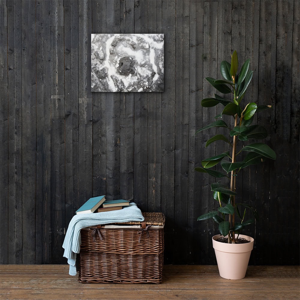 Una cesta de mimbre con libros y una manta doblada reposa sobre un suelo de madera contra una pared revestida de madera oscura, representando el Arte Decorativo. A la derecha se encuentra una planta en maceta con hojas verdes brillantes, que da vida a la esencia de Paisajes Canarios. Arriba, la elegante pintura abstracta monocromática "Lienzo roca y sal" cuelga elegantemente en la pared.