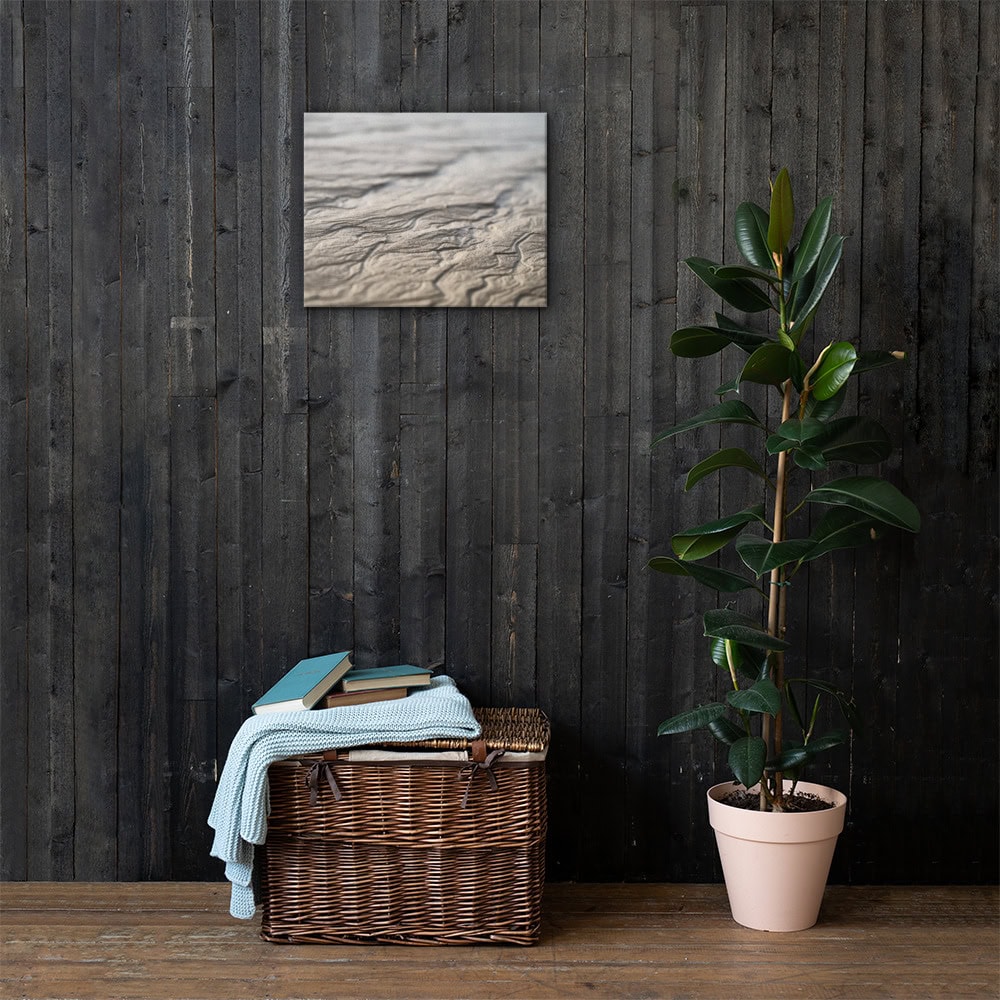 Una cesta de mimbre con libros y una manta doblada reposa contra una pared de madera oscura, lo que añade un toque de Arte Decorativo. Encima cuelga "Lienzo surcos arena mojada", una pieza abstracta que añade intriga visual. Cerca, a la derecha, se encuentra una planta de caucho en una maceta de color rosa pálido, que evoca la esencia serena de La Palma.