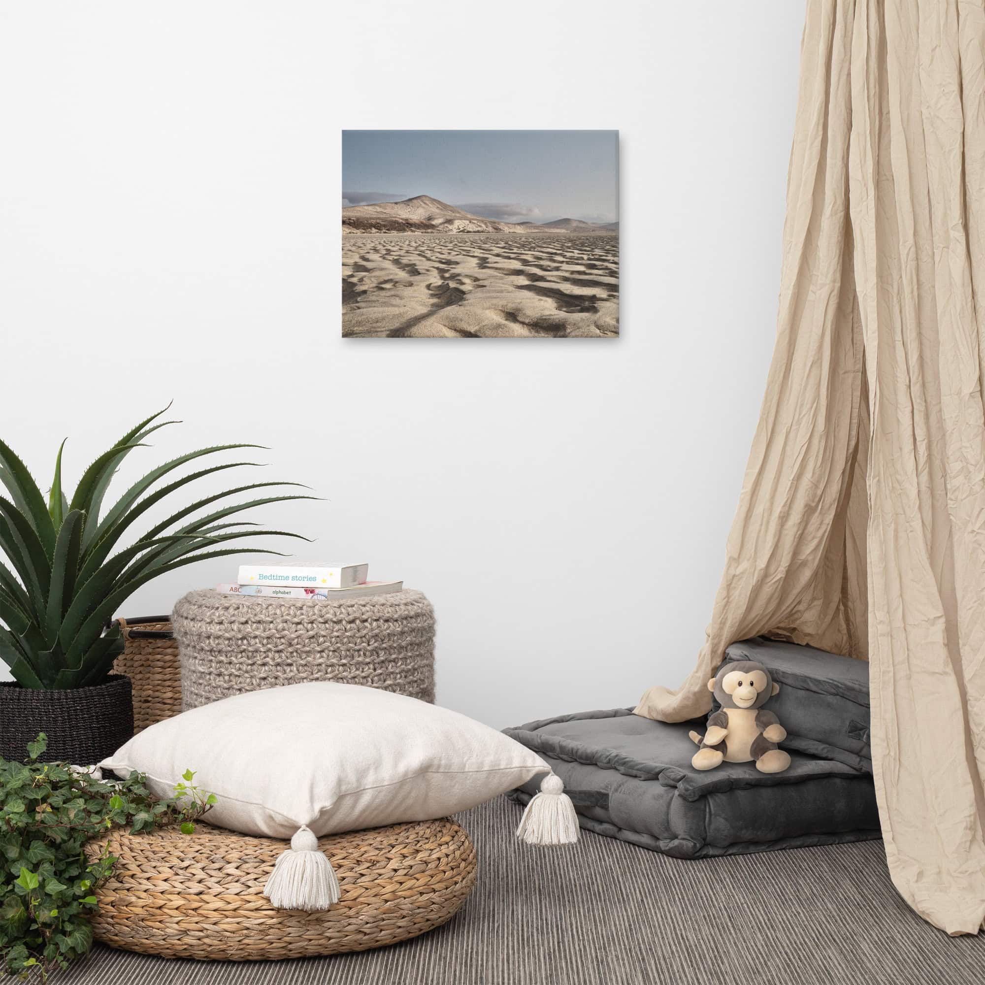 Un rincón acogedor con cojines y una mesita auxiliar rodeada de plantas. Un dosel beige cubre un lateral y una cesta redonda sostiene un mono de peluche. Una fotografía titulada "Lienzo Sotavento", que recuerda a las dunas de arena del desierto, cuelga de la pared blanca.