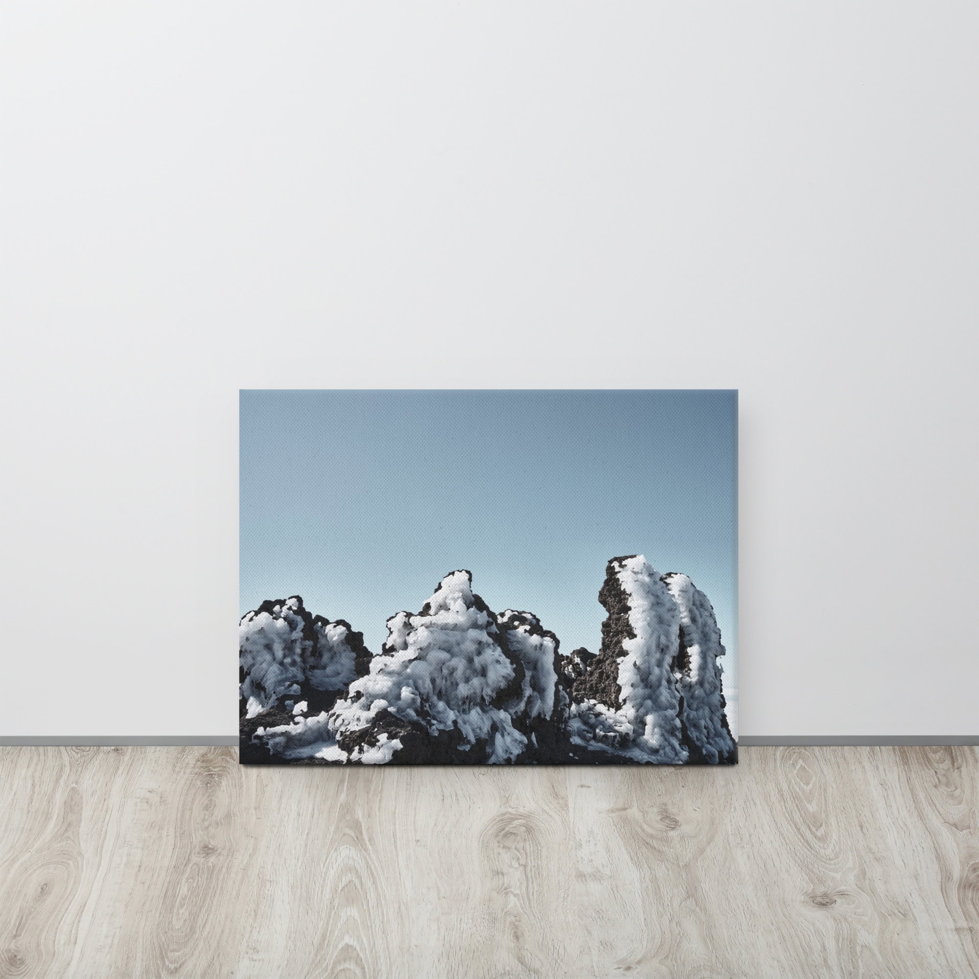 Una fotografía titulada "Lienzo Roque helado" se apoya contra una pared blanca y muestra rocas cubiertas de nieve bajo un cielo azul claro. Esta pieza de arte decorativo reposa sobre un piso de madera, lo que ofrece un contraste con los tonos fríos de la foto.