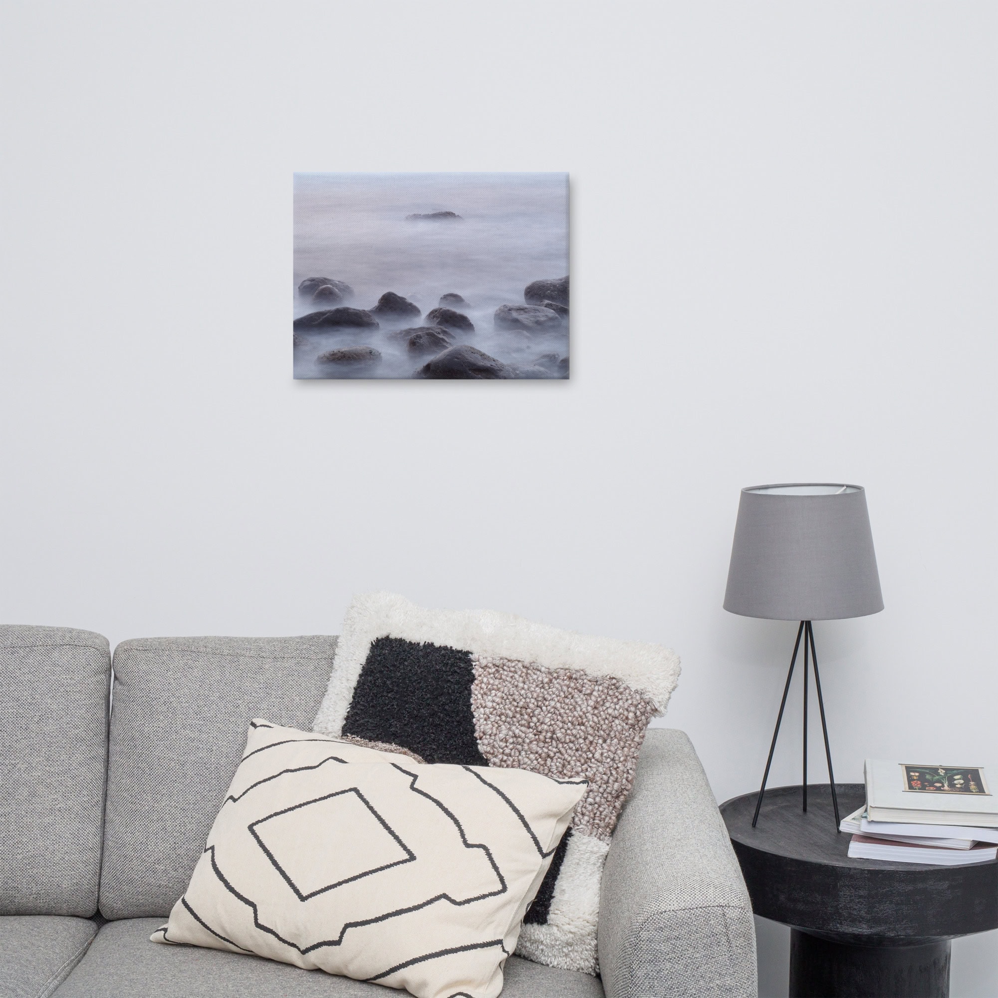 Una sala de estar minimalista cuenta con un sofá gris con cojines decorativos, una mesa auxiliar redonda negra y una pantalla de lámpara gris. El Lienzo de Rocas de la playa de El Lloret está colgado en la pared blanca, lo que agrega un toque de serenidad al espacio.