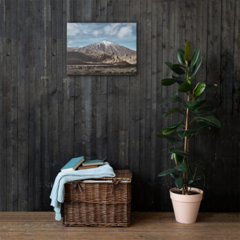 Una habitación rústica con una pared de madera oscura presenta una exquisita pintura de paisaje de una montaña. Debajo, una canasta de mimbre contiene libros y una manta doblada. Una planta alta en maceta se encuentra cerca, agregando vegetación. Para una decoración similar, visite nuestra tienda en línea para obtener impresionantes fotografías impresas del Lienzo Teide.