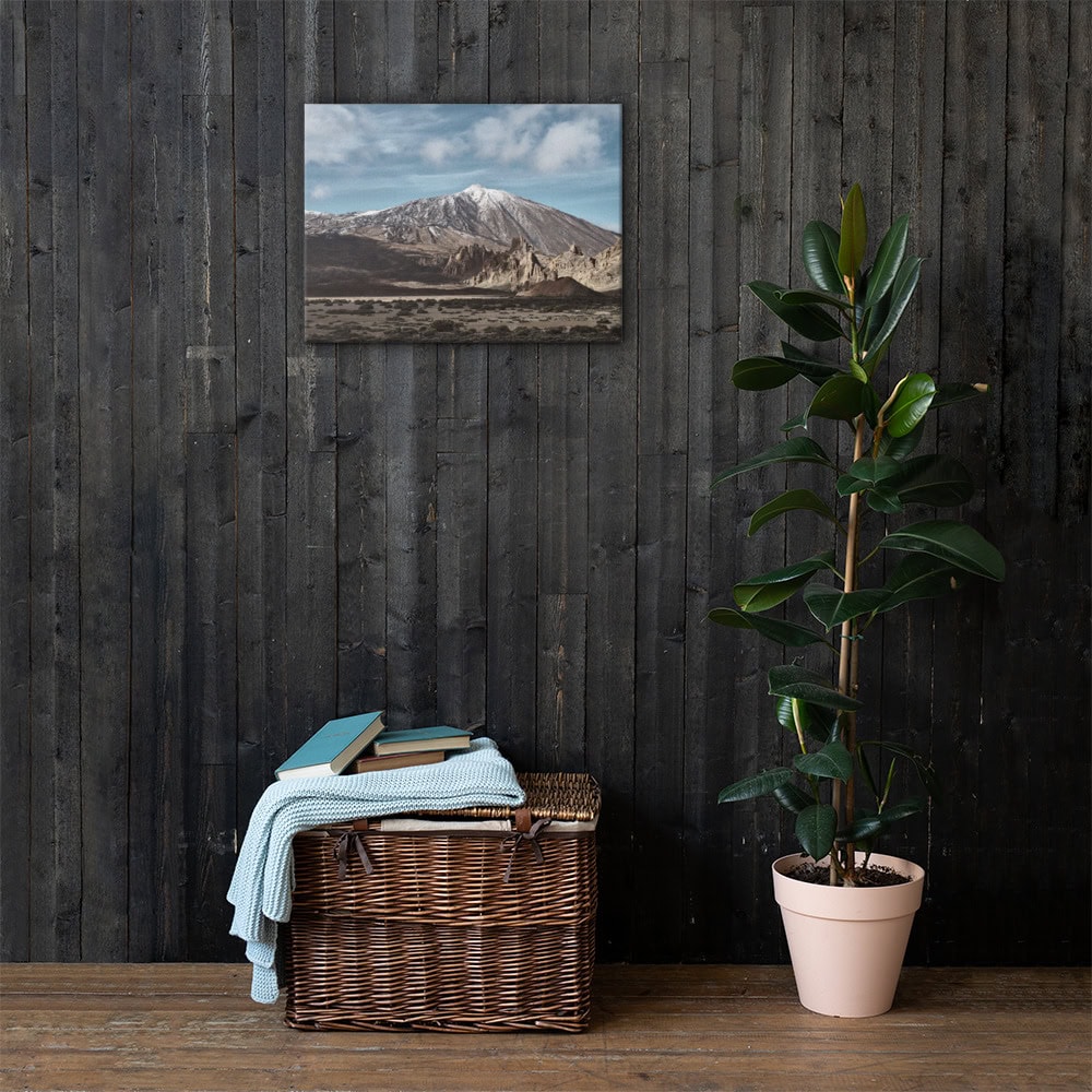 Una habitación rústica con una pared de madera oscura presenta una exquisita pintura de paisaje de una montaña. Debajo, una canasta de mimbre contiene libros y una manta doblada. Una planta alta en maceta se encuentra cerca, agregando vegetación. Para una decoración similar, visite nuestra tienda en línea para obtener impresionantes fotografías impresas del Lienzo Teide.