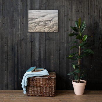 Una canasta de mimbre llena de mantas dobladas y libros reposa sobre un piso de madera contra una pared revestida con paneles de madera oscura. Una planta en maceta con hojas verdes brillantes realza el ambiente acogedor. Arriba, la obra de arte abstracta "Lienzo Surcos Arena Mojada" agrega un toque de arte decorativo que eleva el encanto del espacio.