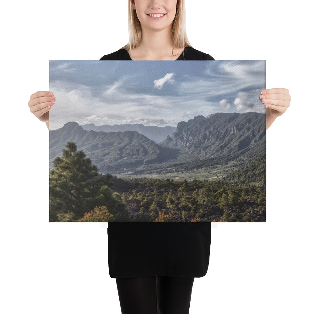Una persona vestida de negro sostiene un cartel que muestra el pintoresco paisaje montañoso de La Palma, adornado con una exuberante vegetación, picos escarpados y un cielo parcialmente nublado. Explore más vistas impresionantes como esta con nuestro "Lienzo Caldera de Taburiente" disponible en nuestra tienda en línea que ofrece exquisitas fotografías impresas.