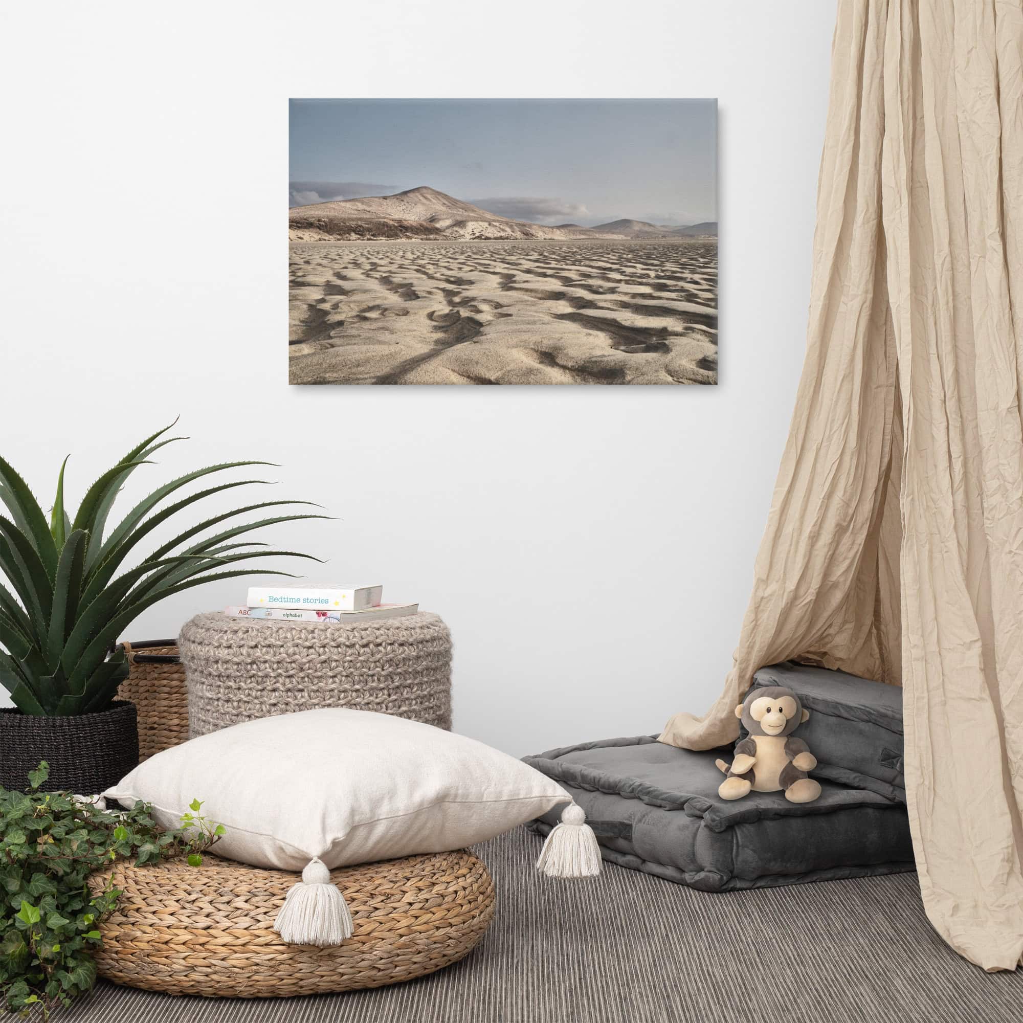 Un rincón acogedor presenta una pequeña pila de cojines grises junto a una cortina beige de Tienda Online. Un simpático mono de peluche reposa sobre un cojín, mientras que una canasta tejida y una planta aportan textura. En la pared blanca cuelga una fotografía de Arte Decorativo del paisaje desértico titulada "Lienzo Sotavento". En el suelo hay cojines de color neutro.