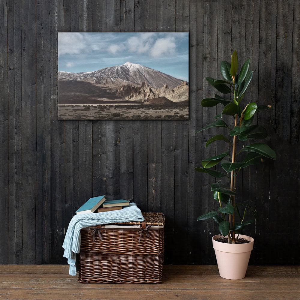 Un interior acogedor con una pared de madera que muestra el paisaje montañoso del Lienzo Teide crea un ambiente relajante. En el suelo, una cesta de mimbre llena de libros y cubierta con una manta azul reposa junto a una planta en maceta con hojas verdes anchas, lo que contribuye a la atmósfera serena.