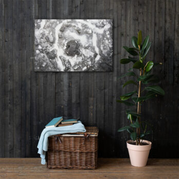 Una cesta de mimbre con libros y una manta azul doblada reposa en el suelo contra una pared de madera oscura. Junto a ella hay una planta en maceta que realza el ambiente acogedor. Arriba, "Lienzo roca y sal", una exquisita pieza de arte decorativo en blanco y negro, está colgada en la pared.