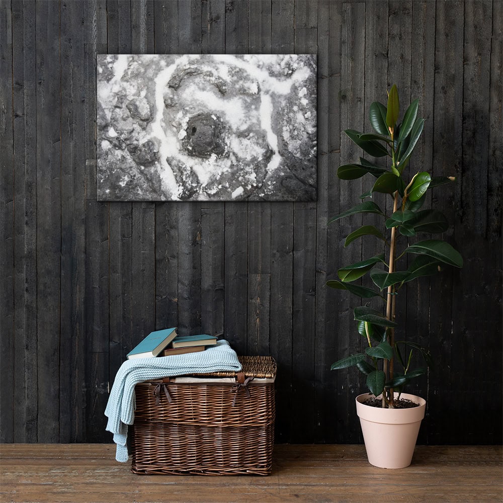 Una cesta de mimbre con libros y una manta azul doblada reposa en el suelo contra una pared de madera oscura. Junto a ella hay una planta en maceta que realza el ambiente acogedor. Arriba, "Lienzo roca y sal", una exquisita pieza de arte decorativo en blanco y negro, está colgada en la pared.