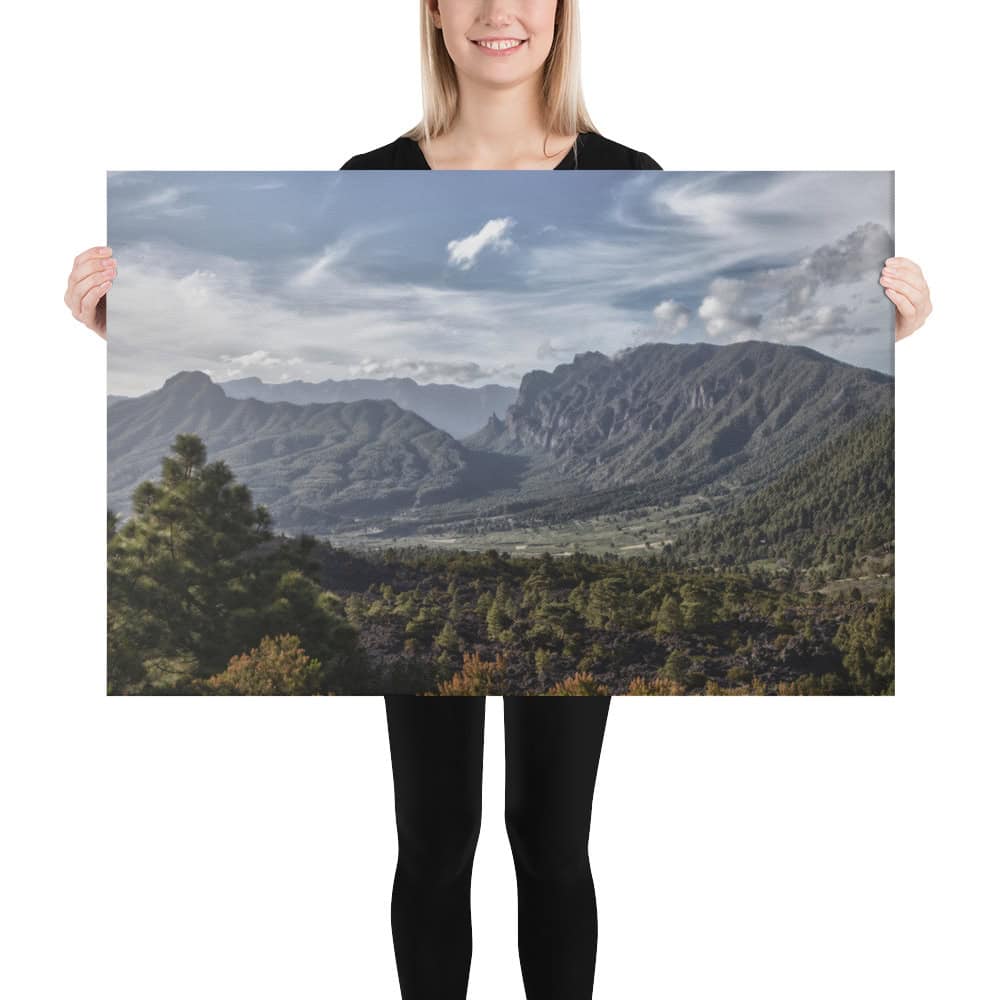 Una persona de Volcanica Productions sostiene un gran cartel de "Lienzo Caldera de Taburiente" que muestra un pintoresco paisaje montañoso con frondosos árboles verdes, colinas onduladas y montañas imponentes bajo un cielo parcialmente nublado. La persona sonríe y viste un atuendo negro, ideal para disfrutar de las impresionantes vistas de La Palma.