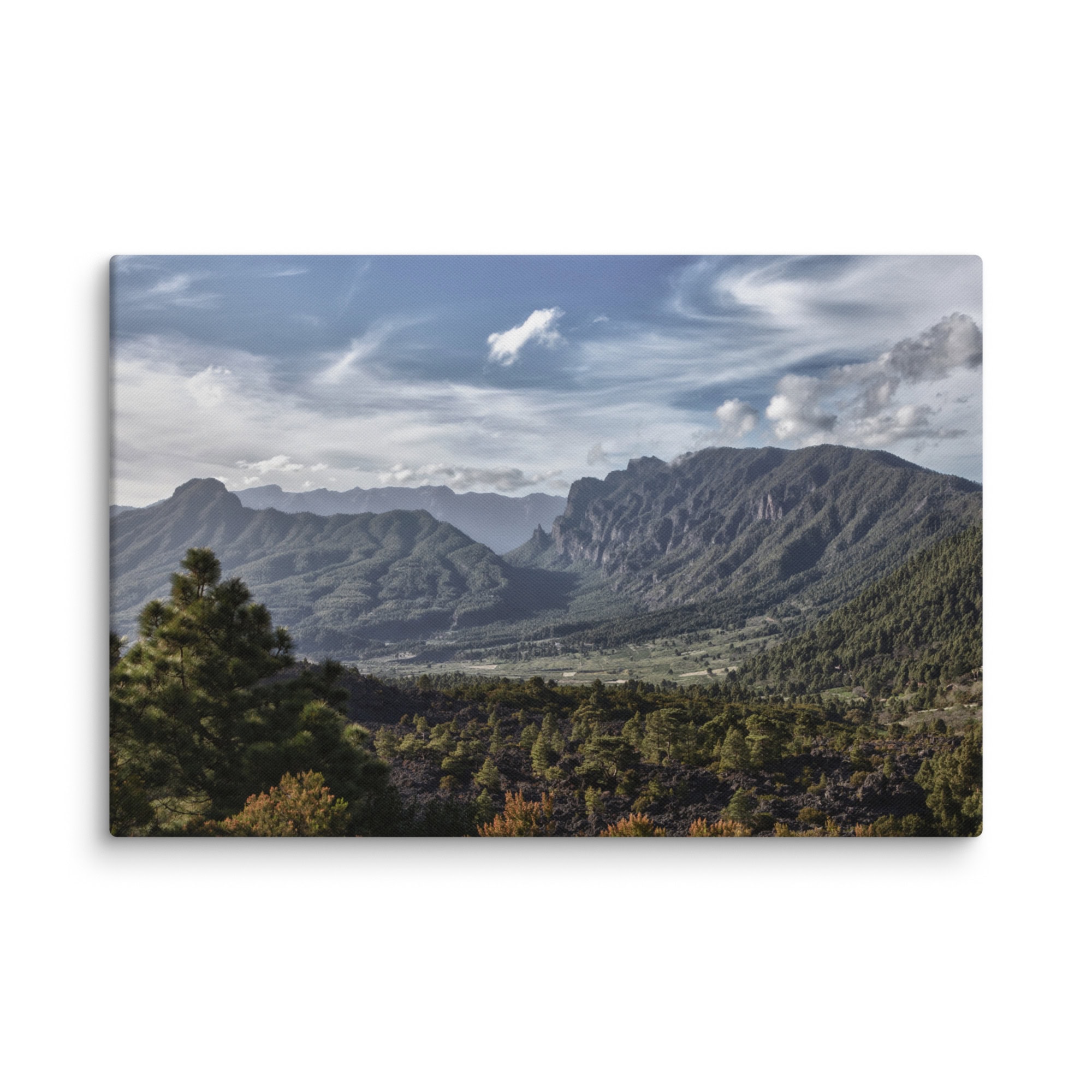 Volcanica Productions capta magistralmente una vista panorámica de un exuberante valle verde rodeado de imponentes montañas bajo un cielo azul parcialmente nublado. Los pinos adornan el primer plano, con nubes dispersas sobre este impresionante paisaje de La Palma, disponible como "Lienzo Caldera de Taburiente" fotografías impresas.