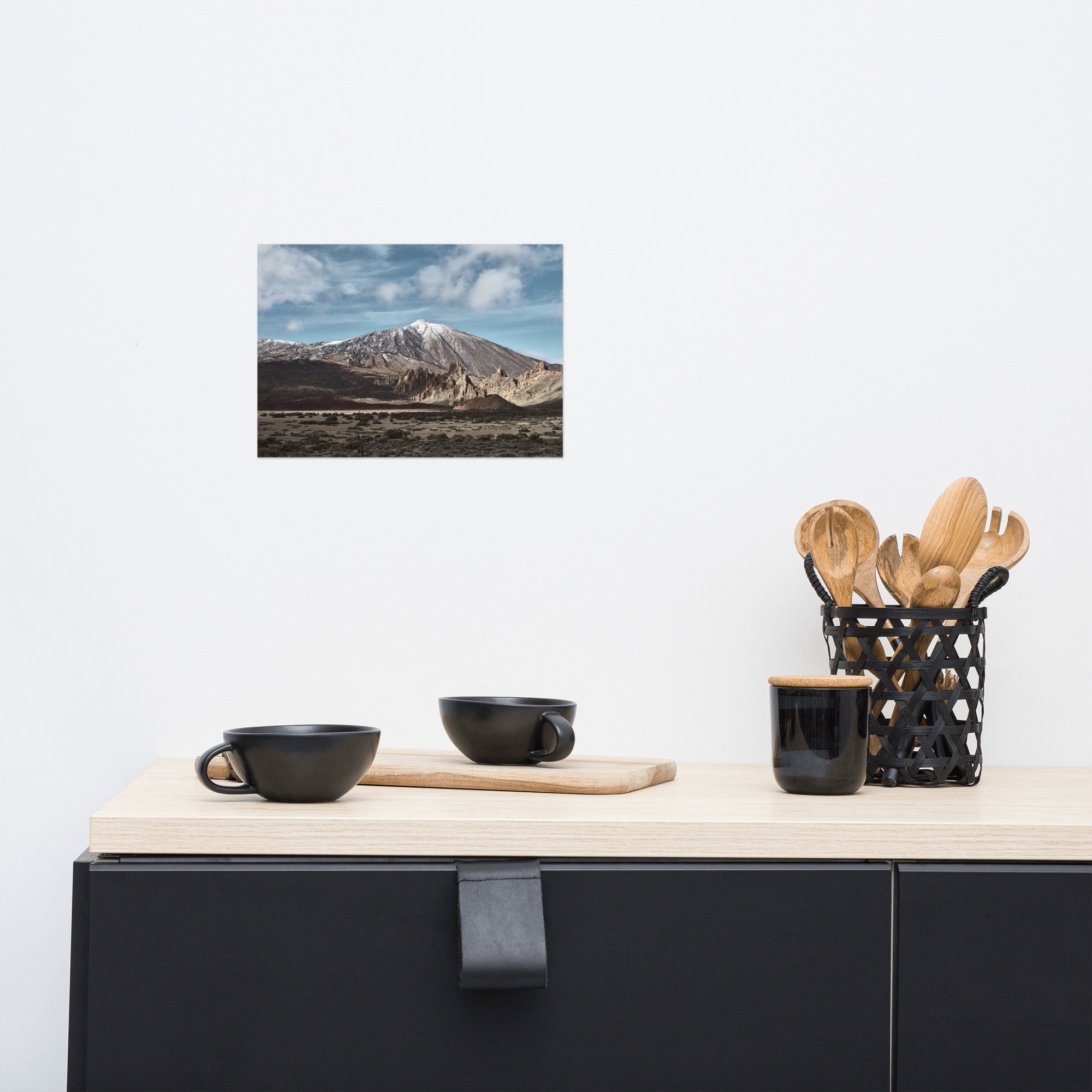 Una encimera de cocina minimalista presenta tazas negras sobre un platillo de madera con un soporte negro para utensilios de madera. Arriba, el cartel "Teide" de Volcanica Productions muestra una montaña nevada bajo un cielo nublado, centrada en una pared blanca.