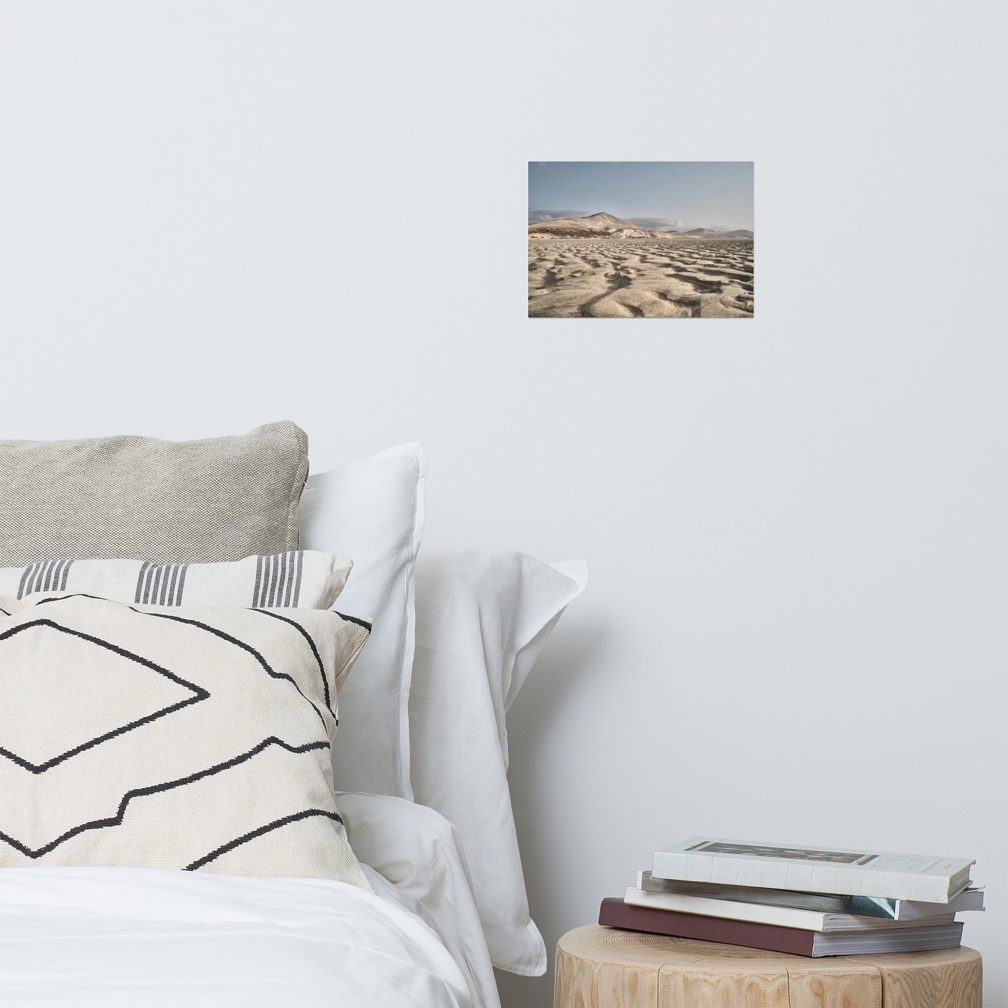 Un dormitorio minimalista cuenta con una cama cuidadosamente tendida con almohadas blancas y estampadas. Una pequeña pila de libros reposa sobre una mesa auxiliar redonda de madera de Tienda Online. Sobre la cama, el cartel "Sotavento" adorna la pared blanca nítida, lo que le da un toque aventurero.