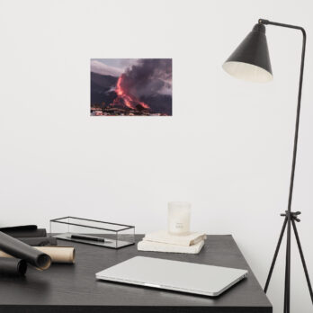 El espacio de trabajo minimalista muestra un escritorio negro adornado con una computadora portátil cerrada, papeles enrollados, una vela y una caja. A la derecha hay una elegante lámpara negra, mientras que arriba, el "Póster volcán 2021 La Palma edificios" de Volcanica Productions muestra un volcán en erupción en La Palma contra la pared blanca prístina.