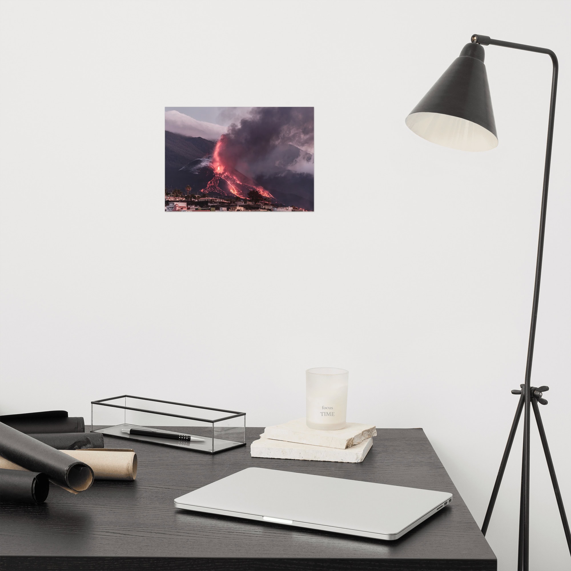 El espacio de trabajo minimalista muestra un escritorio negro adornado con una computadora portátil cerrada, papeles enrollados, una vela y una caja. A la derecha hay una elegante lámpara negra, mientras que arriba, el "Póster volcán 2021 La Palma edificios" de Volcanica Productions muestra un volcán en erupción en La Palma contra la pared blanca prístina.