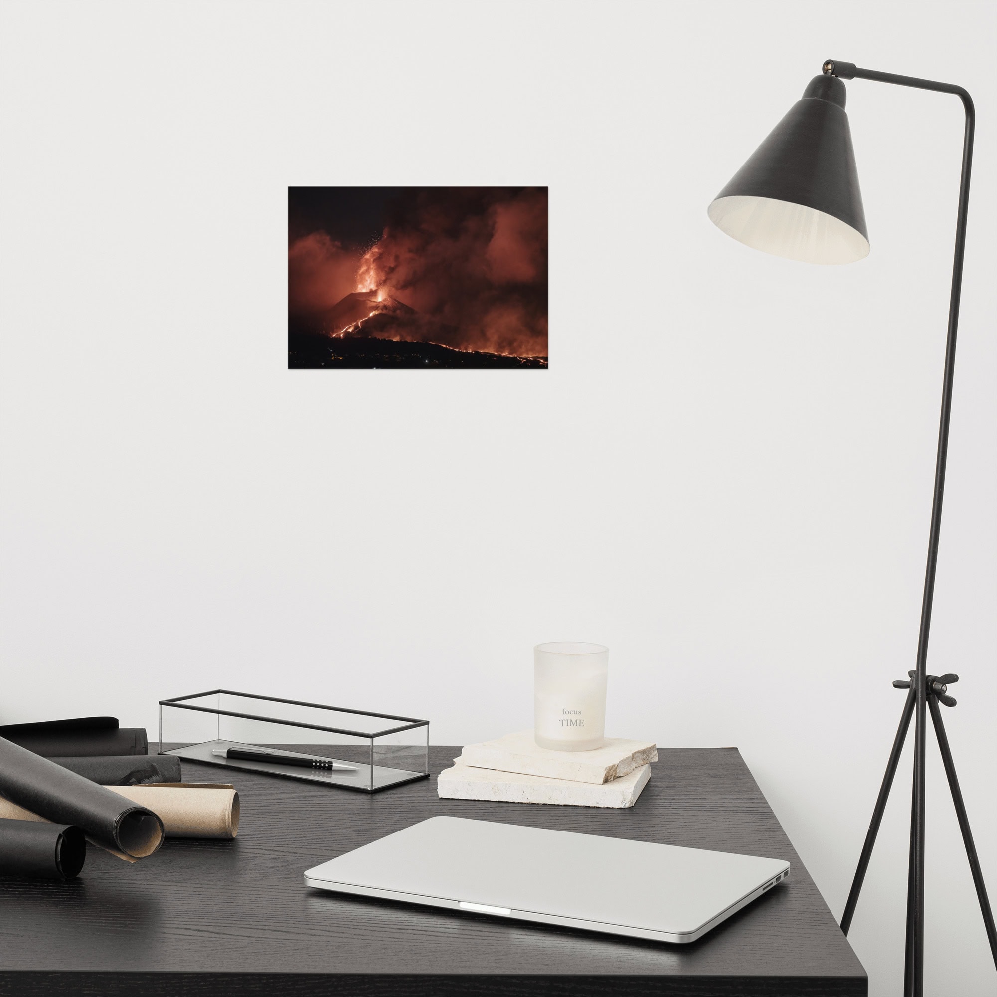 Un espacio de trabajo minimalista muestra un escritorio negro con una computadora portátil cerrada, una vela y libros ordenados ordenadamente. Una lámpara negra moderna se encuentra al lado del escritorio, mientras que la pared de arriba está adornada con una impresionante fotografía titulada "Póster volcán 2021 La Palma noche" de Volcanica Productions, que muestra un volcán en erupción.