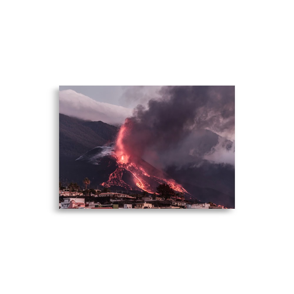 Un volcán entra en erupción y cae lava de color naranja vibrante por sus laderas, mientras un denso humo negro se arremolina en el cielo. Debajo, un pequeño pueblo descansa bajo un cielo nublado, encapsulando la espectacular belleza de la escena capturada en el "Póster volcán 2021 La Palma edificios", ideal para fotografías impresionantes.