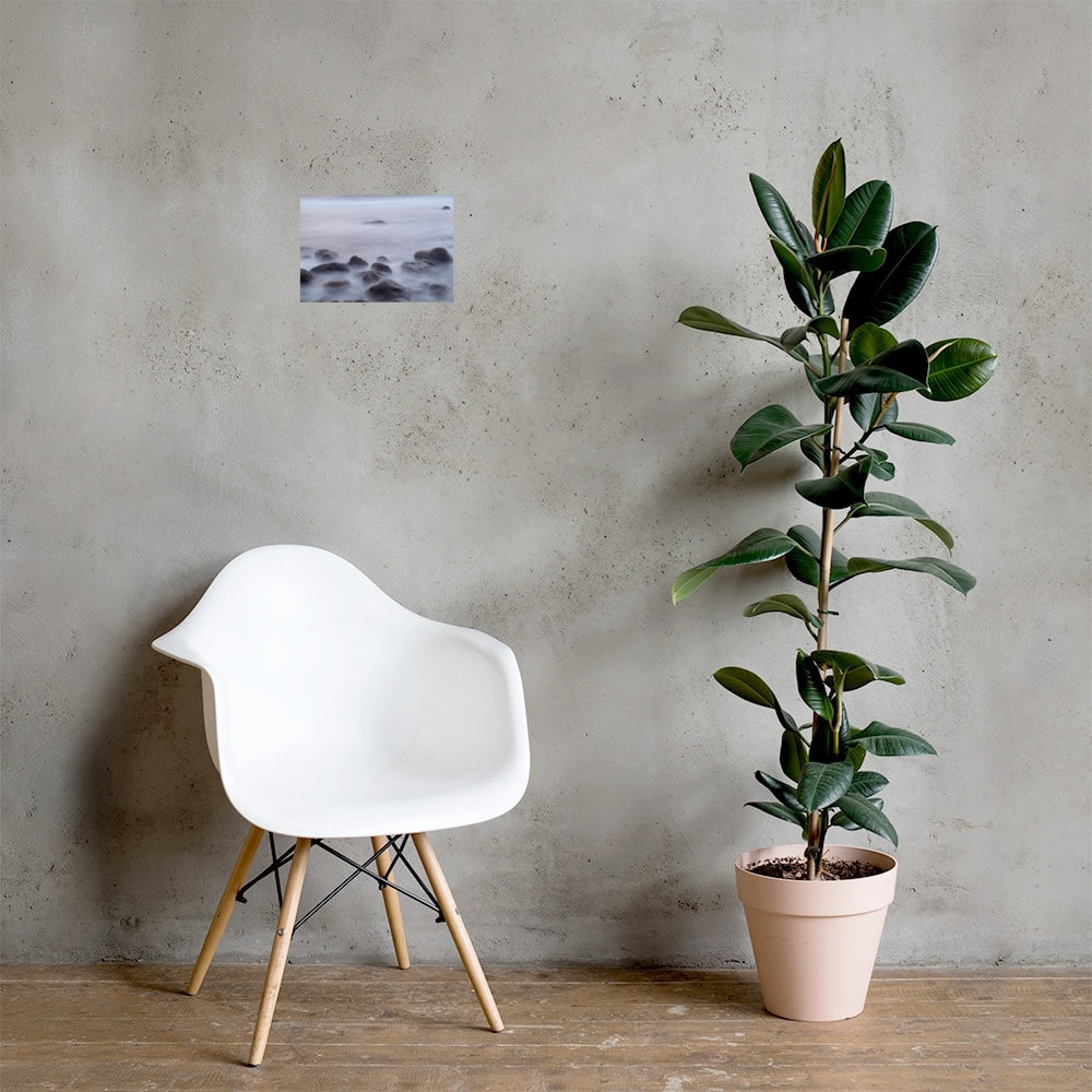 Una moderna silla blanca con patas de madera se encuentra junto a una maceta con una planta verde alta sobre un suelo de madera, que recuerda a la serenidad de Paisajes Canarios. Al fondo, el cartel "Póster de Rocas de la playa de El Lloret" cuelga de una pared gris lisa, lo que añade un aire artístico inspirado en la belleza de La Palma.