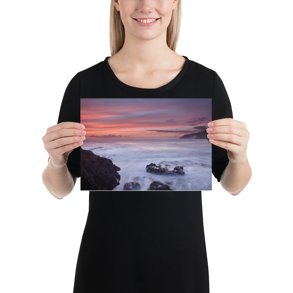 Una persona con una camisa negra sostiene una foto de la impresionante vista del océano al atardecer de Volcanica Productions, titulada "Póster rocas de Los Guirres". El cielo está adornado con tonos de rosa y púrpura mientras las olas chocan suavemente contra las rocas en primer plano; una hermosa pieza disponible en su tienda en línea.