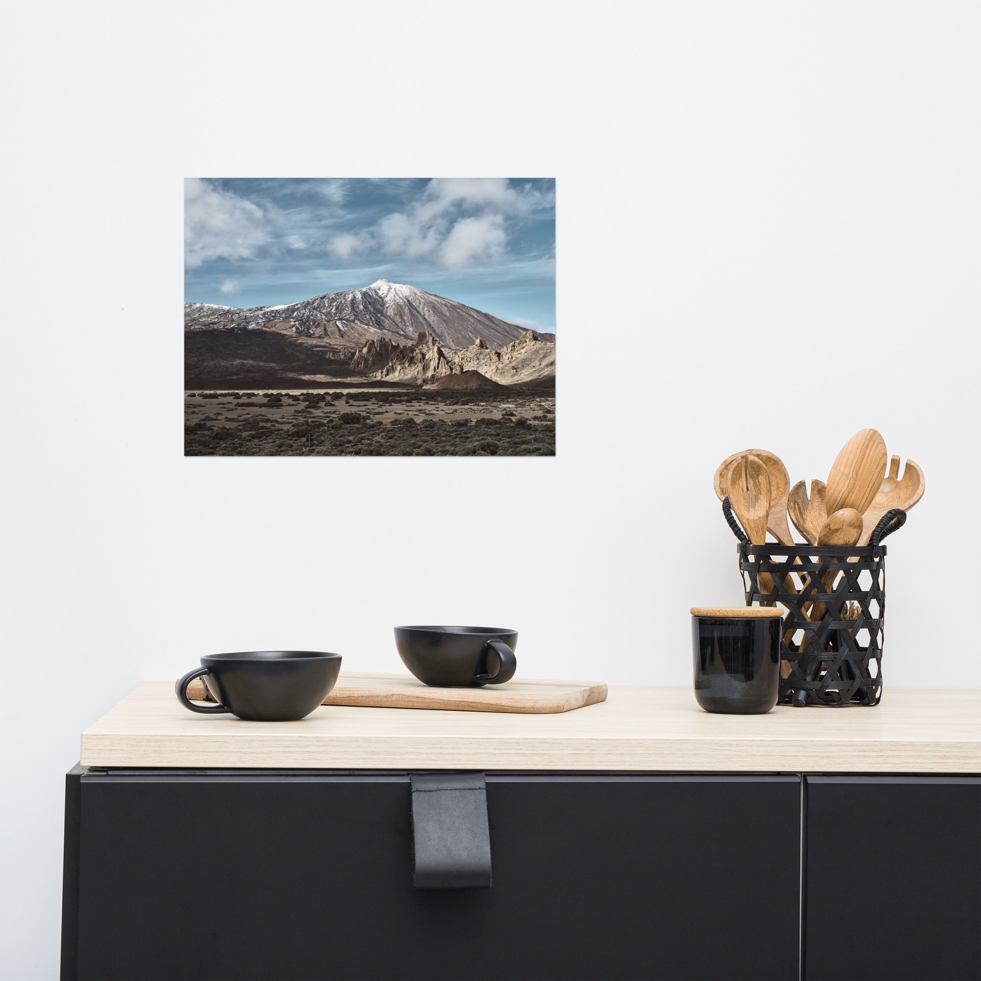 Una moderna encimera de cocina con tazas negras y un portautensilios ocupa el centro del escenario, mientras que una foto de Póster Teide de una montaña cubierta de nieve bajo un cielo nublado adorna la pared blanca.