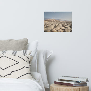 Un acogedor rincón de dormitorio con una cama, almohadas y una mesa auxiliar redonda repleta de libros presenta una fotografía horizontal enmarcada de Póster Sotavento colgada en la pared blanca sobre la cama, agregando un toque exótico a la decoración.