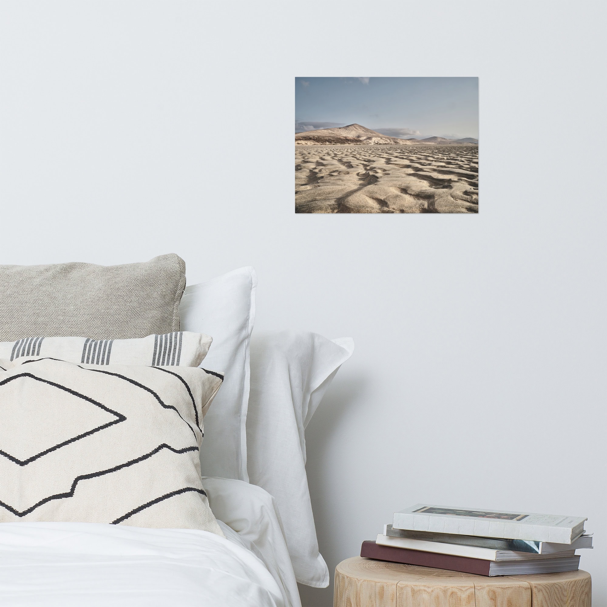 Un acogedor rincón de dormitorio con una cama, almohadas y una mesa auxiliar redonda repleta de libros presenta una fotografía horizontal enmarcada de Póster Sotavento colgada en la pared blanca sobre la cama, agregando un toque exótico a la decoración.