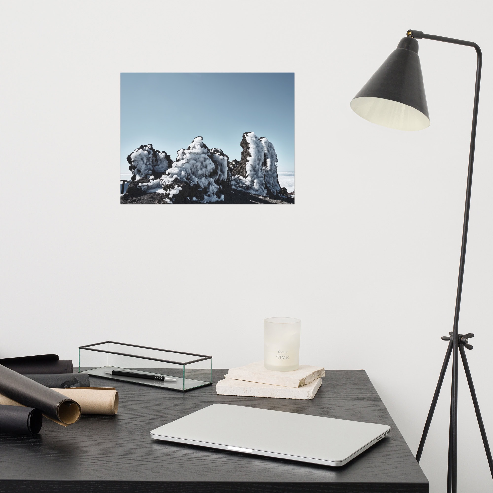 Una oficina minimalista diseñada por Volcanica Productions muestra un escritorio negro adornado con una computadora portátil cerrada, una elegante lámpara de escritorio, una vela y elementos decorativos. En la pared blanca de arriba cuelga el "Póster Roque helado", que representa las formaciones rocosas nevadas de La Palma.