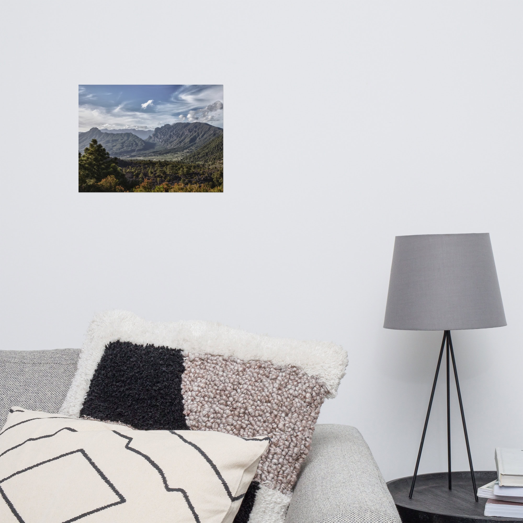 Una moderna sala de estar cuenta con una lámpara gris con soporte de trípode, un sofá adornado con cojines con dibujos geométricos y una pequeña pintura de paisaje titulada "Póster Caldera de Taburiente" que representa montañas y árboles en la pared blanca.