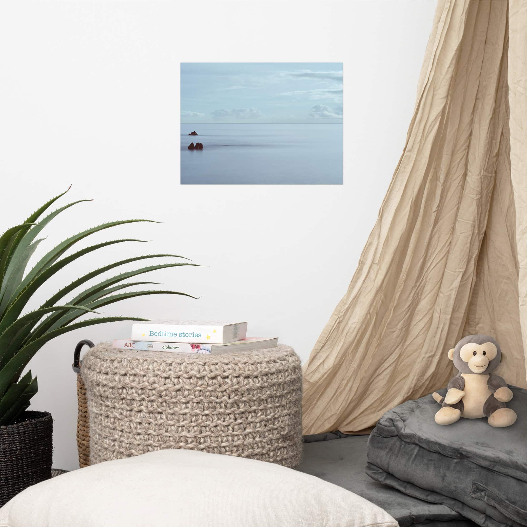 Un rincón acogedor cuenta con un dosel de color beige, un mono de peluche, cojines grises apilados y un puf tejido con libros. Una planta en maceta reposa en el suelo, realzada por el póster Mar en Calma en Los Cancajos que se exhibe en la pared blanca.