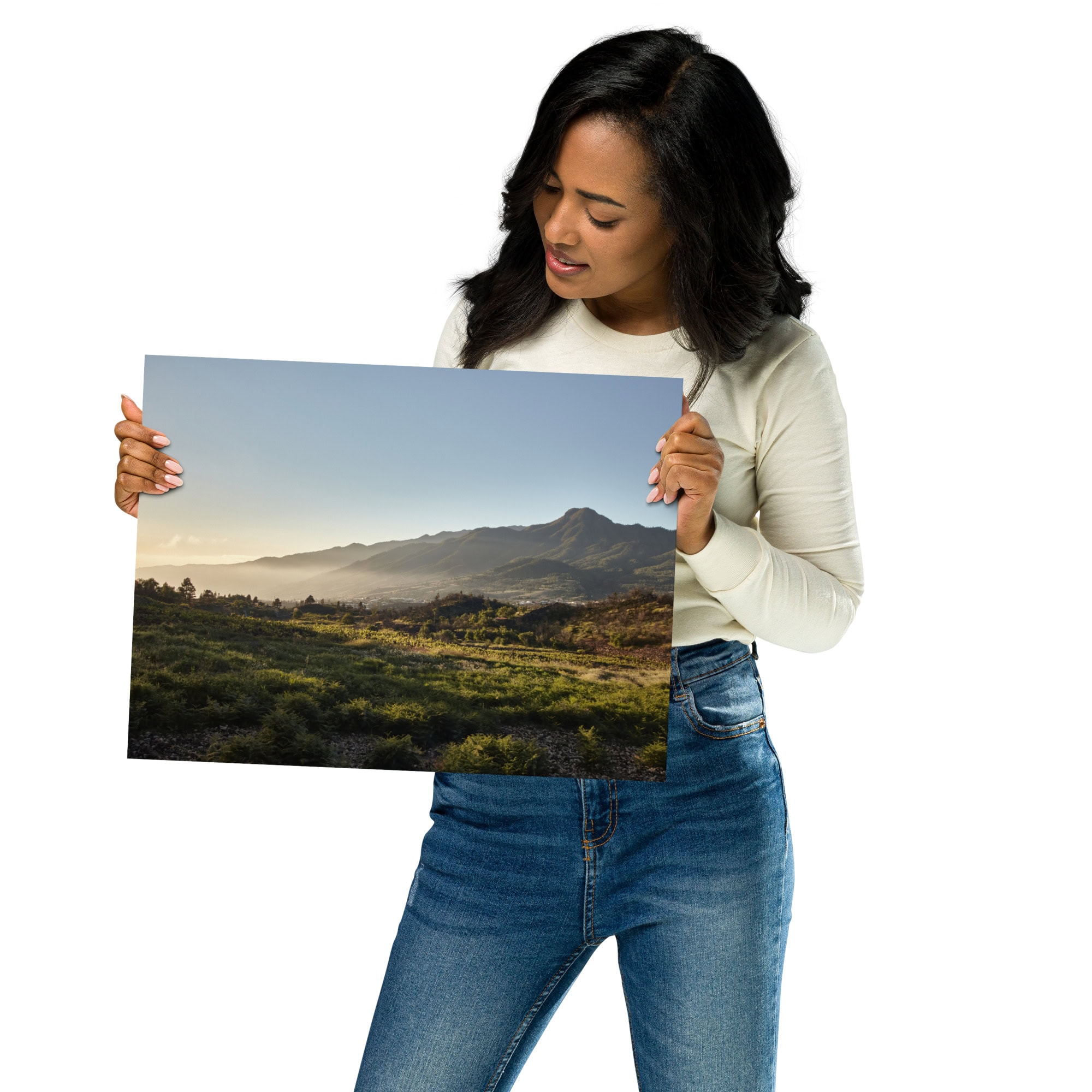 Una mujer con una camisa blanca de manga larga y jeans sostiene una fotografía grande titulada "Póster atardecer desde Cuesta Juliana" de Volcanica Productions, que muestra las majestuosas montañas de La Palma y su exuberante vegetación bajo un cielo despejado.