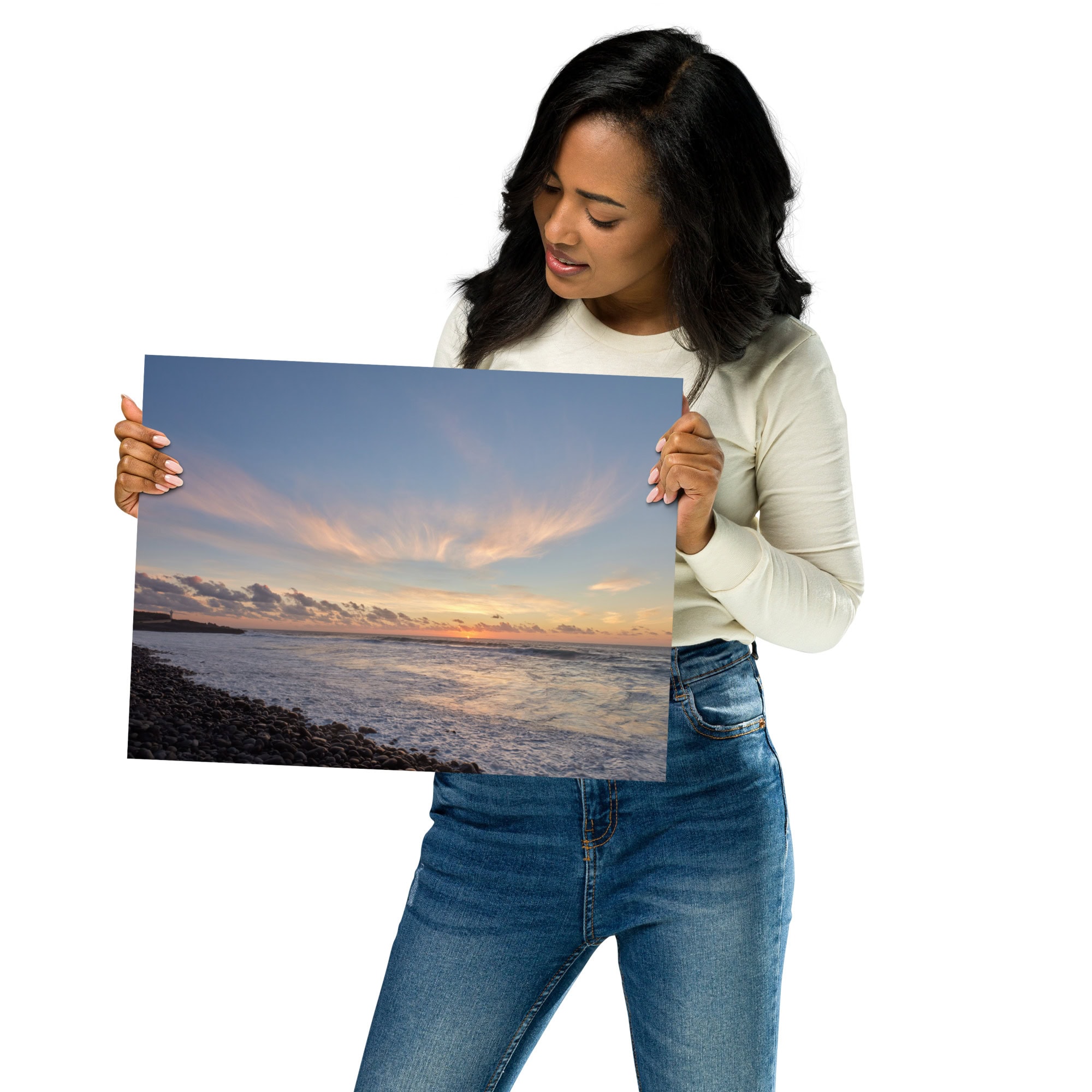 Una mujer con una camisa blanca de manga larga y jeans sostiene una gran fotografía de un paisaje costero al atardecer, titulada "Póster atardecer en Los Guirres" de Volcanica Productions. Esta pieza captura una costa de guijarros, un mar en calma y nubes que reflejan tonos cálidos. La mujer examina el cartel de cerca, apreciando cada detalle.