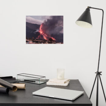 Un escritorio moderno con un ordenador portátil, velas y papeles enrollados se ve realzado por una lámpara negra. En la pared, un "Póster volcán 2021 La Palma edificios" de Paisajes Canarios muestra un volcán en erupción en La Palma, con un humo espeso y lava fluyendo que crean un fondo espectacular.