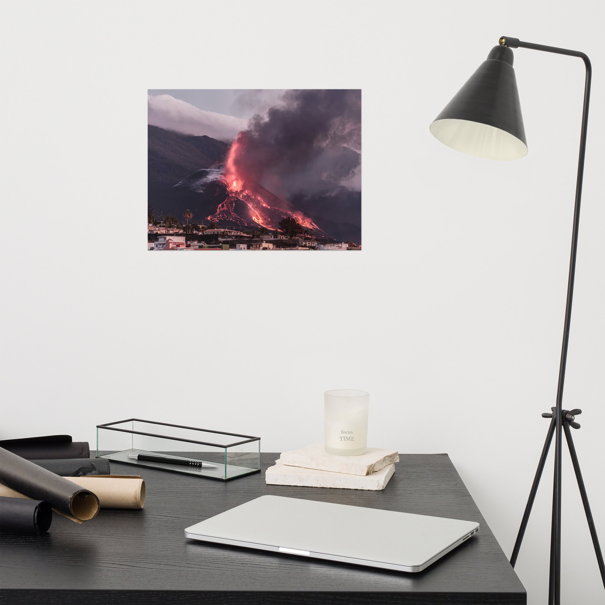 Un escritorio moderno con un ordenador portátil, velas y papeles enrollados se ve realzado por una lámpara negra. En la pared, un "Póster volcán 2021 La Palma edificios" de Paisajes Canarios muestra un volcán en erupción en La Palma, con un humo espeso y lava fluyendo que crean un fondo espectacular.