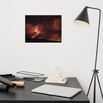 Un espacio de trabajo minimalista muestra un escritorio negro adornado con una computadora portátil cerrada, dos papeles negros enrollados, una vela y un marco de fotos. Una lámpara en blanco y negro está ubicada al lado del escritorio. En la pared cuelga un impresionante Póster volcán 2021 La Palma noche, que captura la ardiente erupción volcánica por la noche y agrega un toque de arte decorativo.